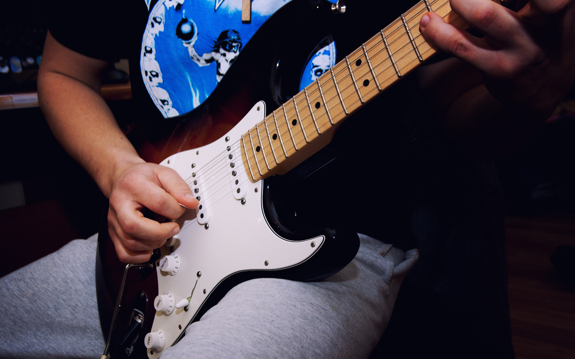 A man in a black T-shirt and gray sweatpants plays a Fender Stratocaster electric guitar.