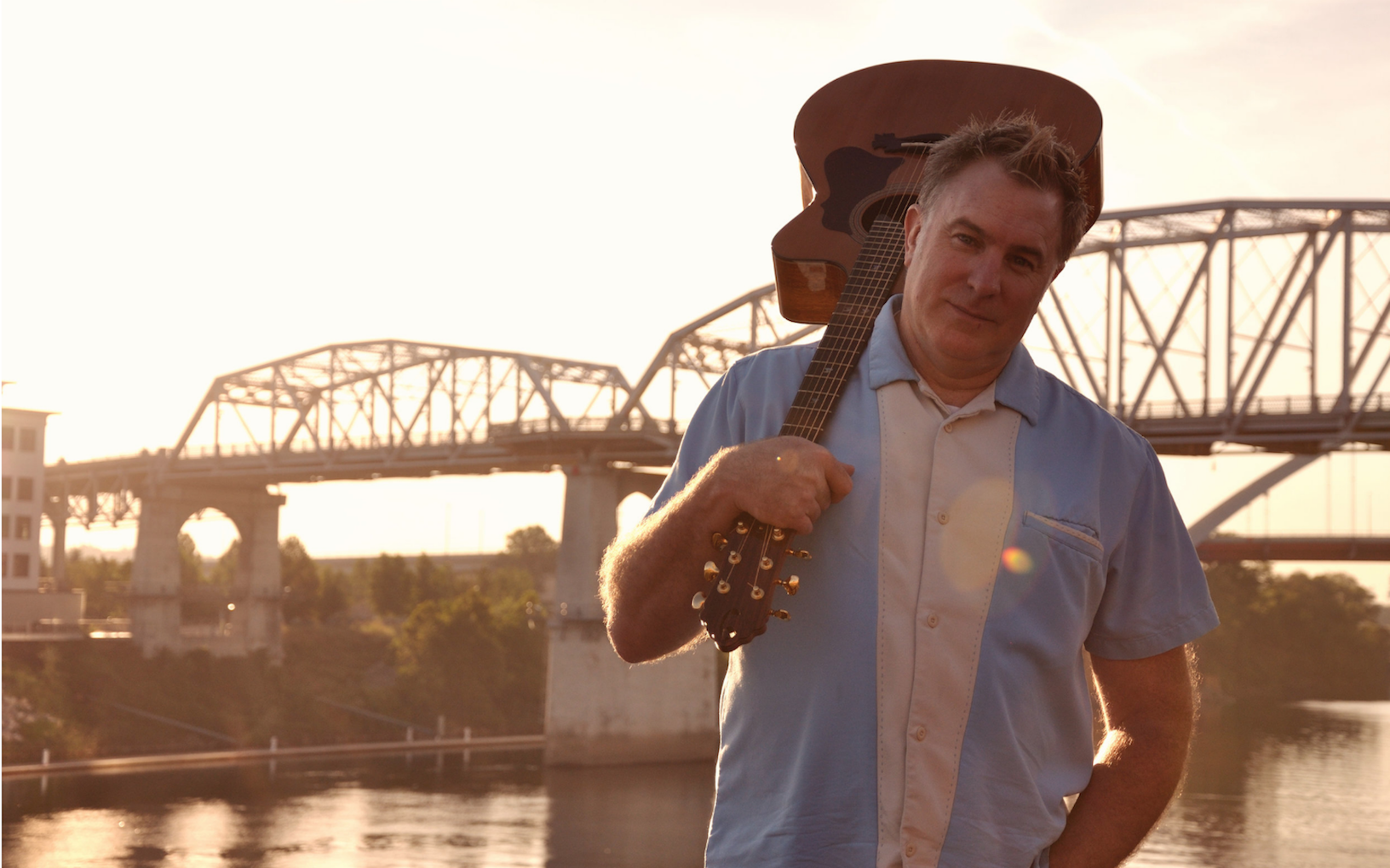 Songwriter Shane Adams with a guitar over his shoulder next to a bridge