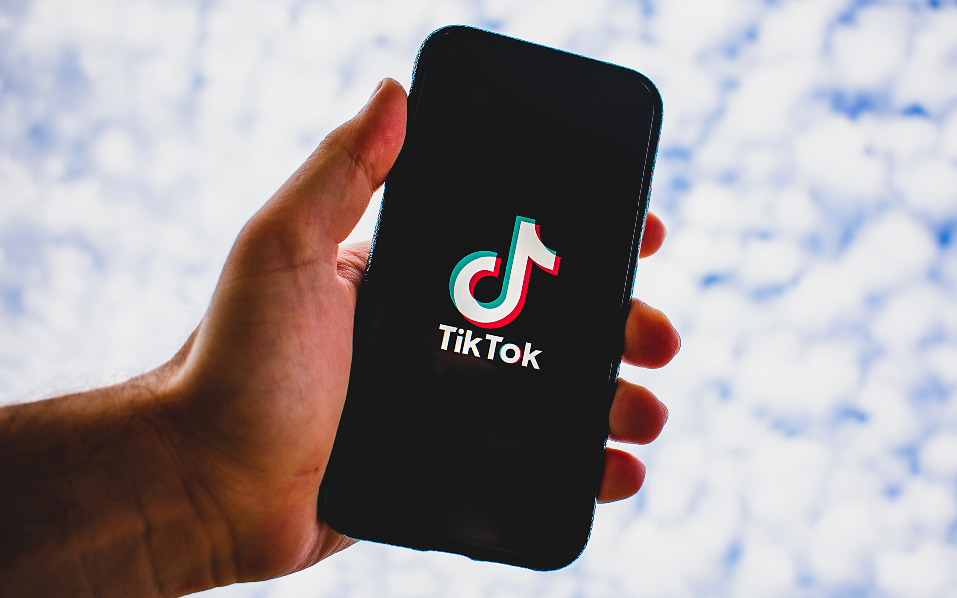 A hand holds a cell phone, featuring the TikTok logo on the screen, towards the clouded sky.  