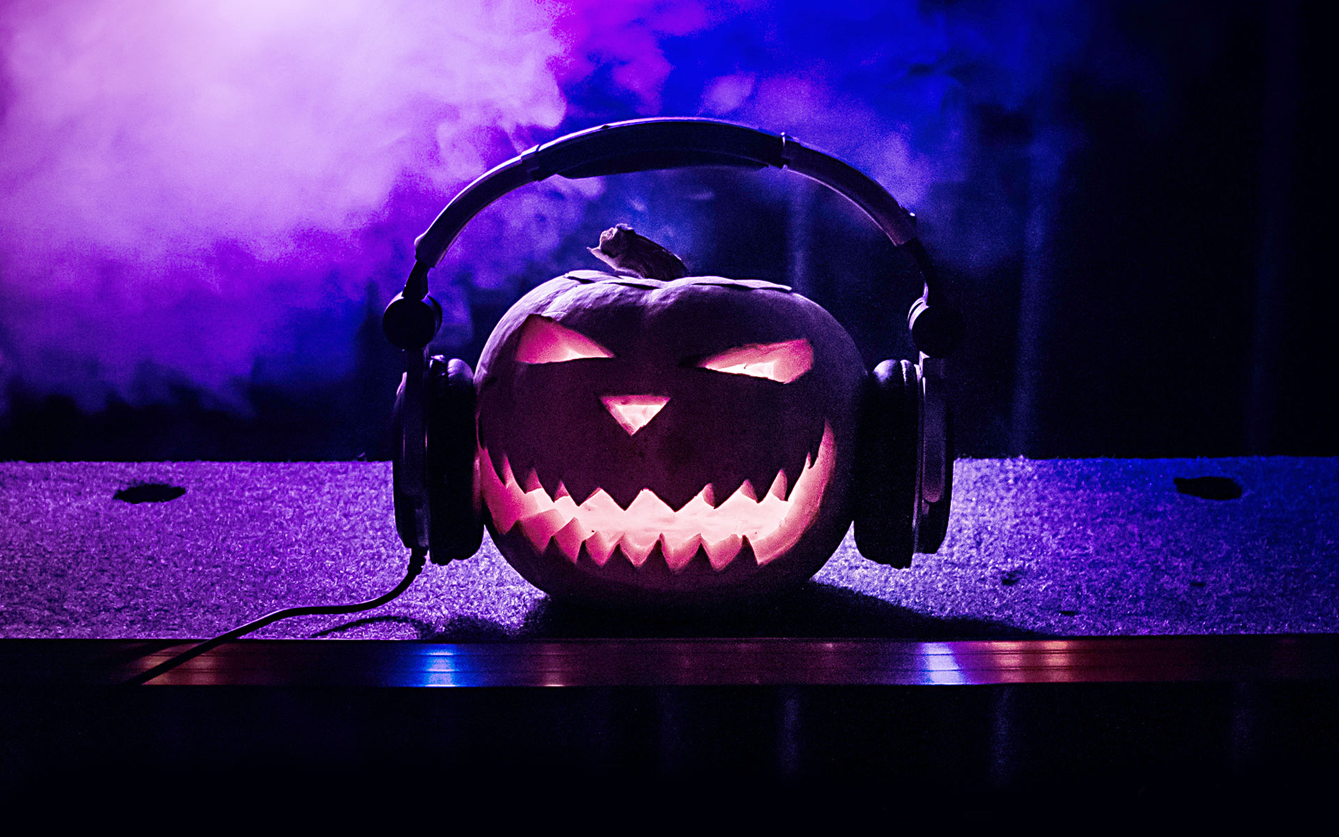 An illuminated jack o' lantern wearing headphones sits on top of a soundboard surrounded by purple smoke, all to celebrate the spooky music of Halloween.