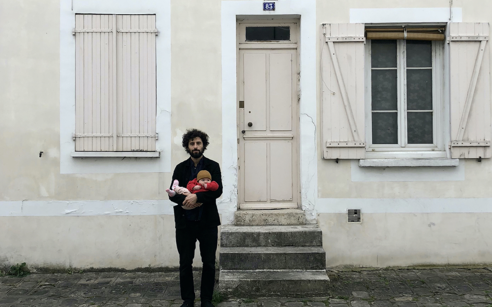 Singer, songwriter, and musician José González is pictured holding a small child while standing outside of a doorway that is marked with the number 83.