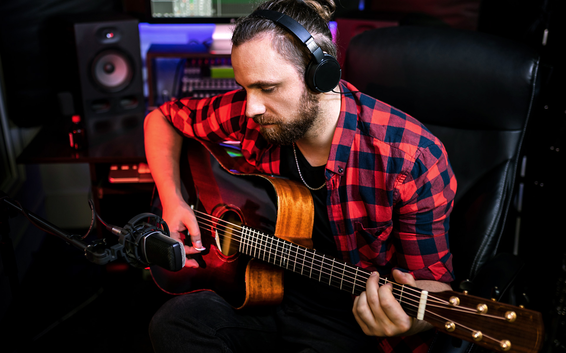 A man with a plaid shirt sits at a mic in a recording studio, presumably to record a guitar part for a song he is hoping to one day land a sync licensing deal with.