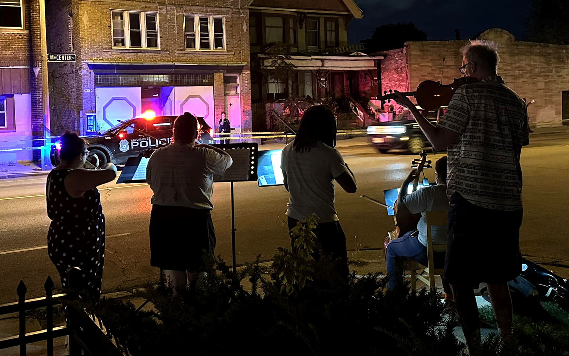 A group of five strings musicians performing at night on the sidewalk next to a police car and "do not cross" tape.