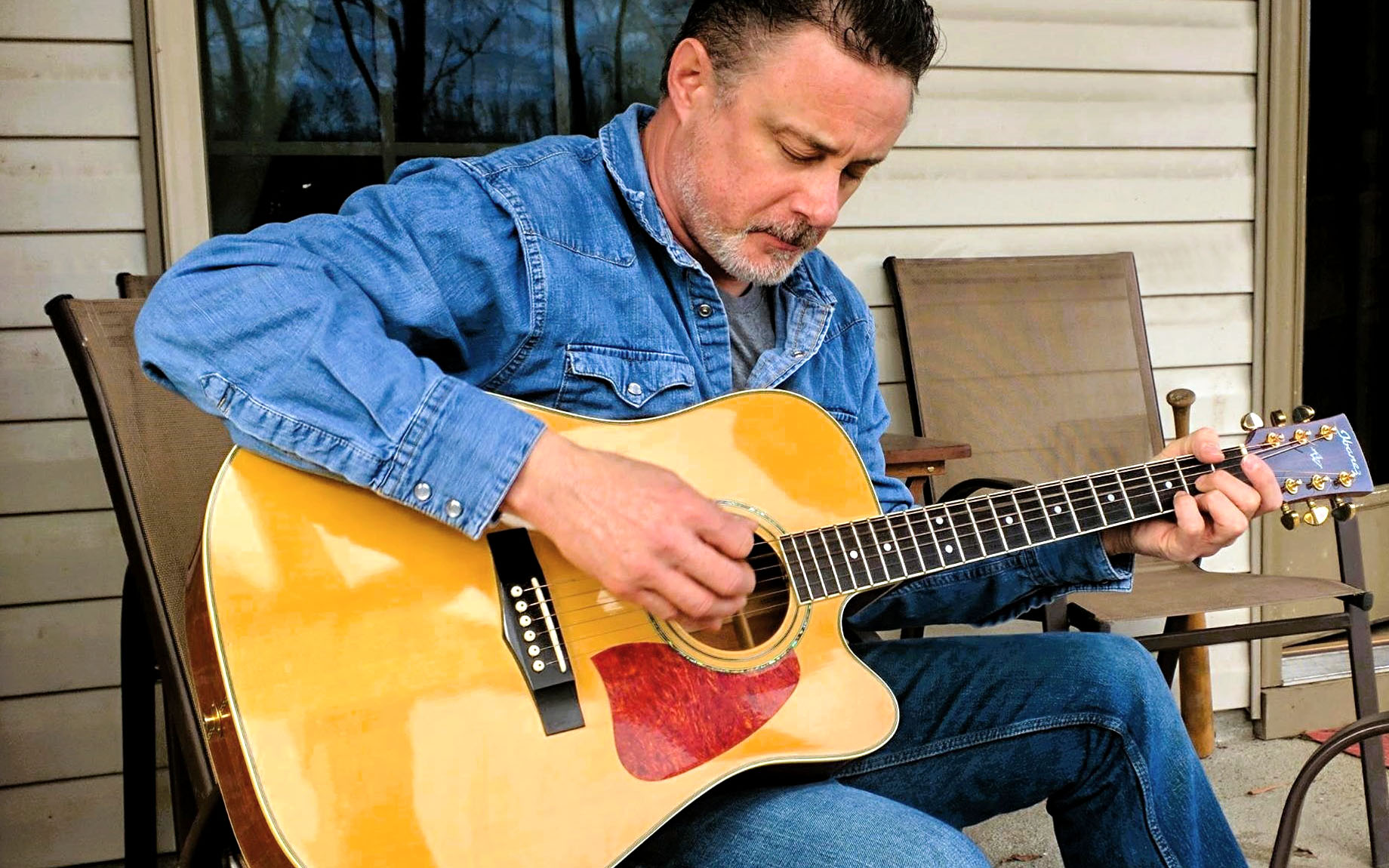 Billy Ray McClelland plays acoustic guitar in a chair in front of a house.