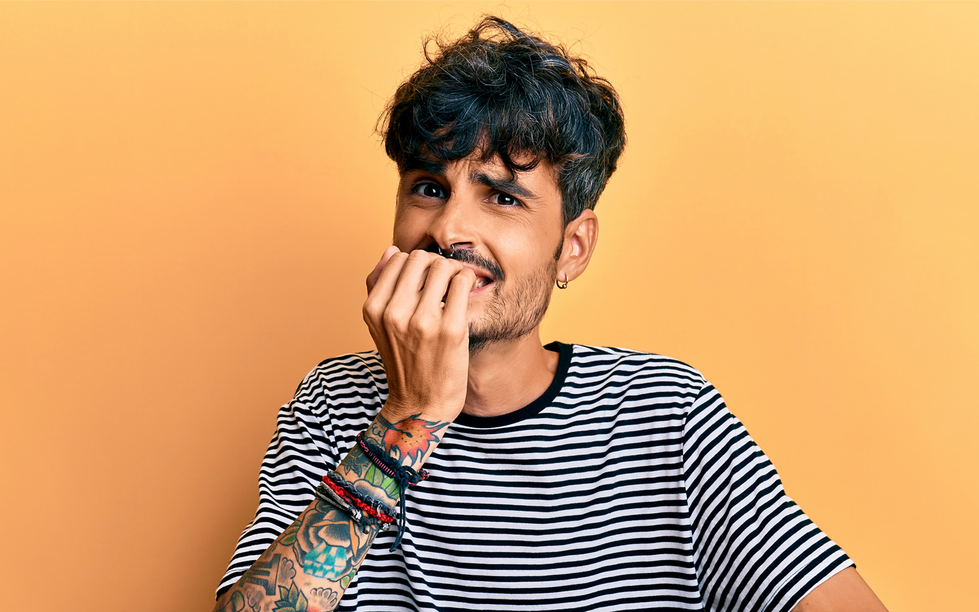 A person with tattoos biting their fingernails and looking nervous against a yellow backdrop. 