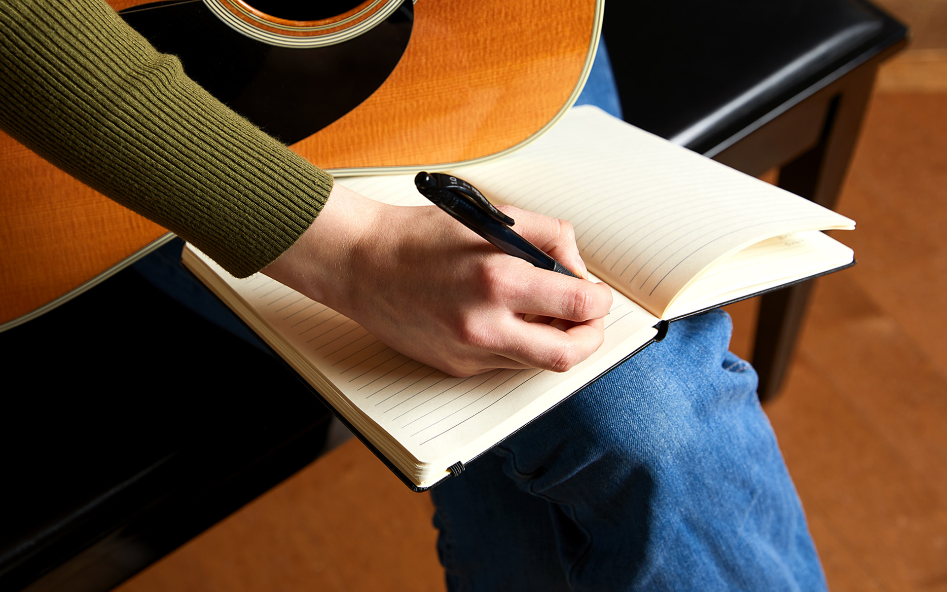 Person's hand writing in a notebook. Guitar on lap in background.