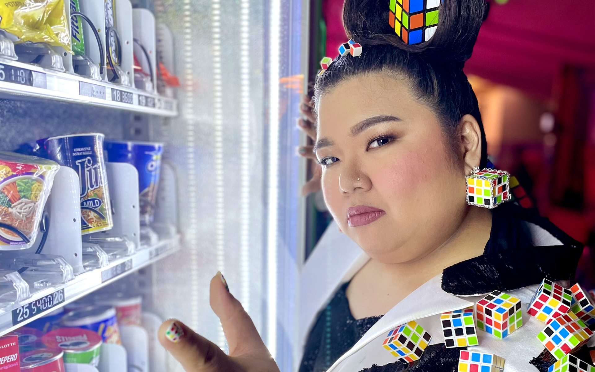 An artistic photo of Ashley Aunor standing in front of a glowing vending machine with rubik's cubes in their hair, on their jacket, and on earrings.  