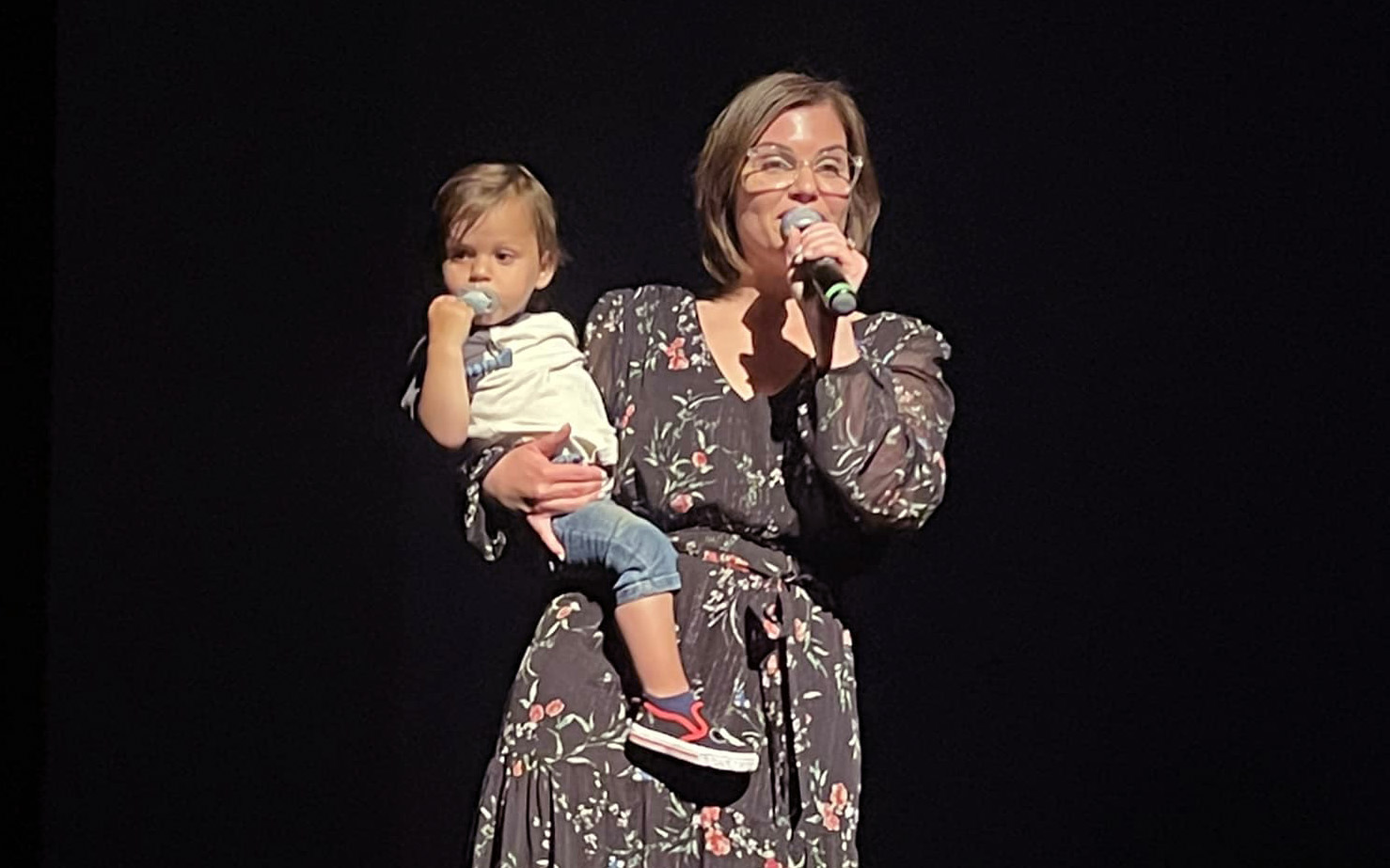 Jaymee holding her then one-year-old son on her hip, with a microphone in the other hand, giving a welcome speech onstage during her production of Mean Girls Jr.