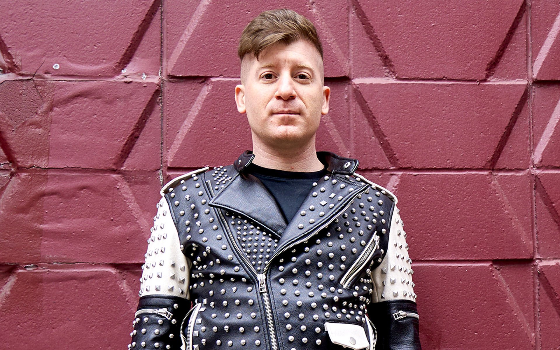 Berklee Online course author and instructor Ben Camp stands against a red brick wall, wearing a black and white studded leather jacket.