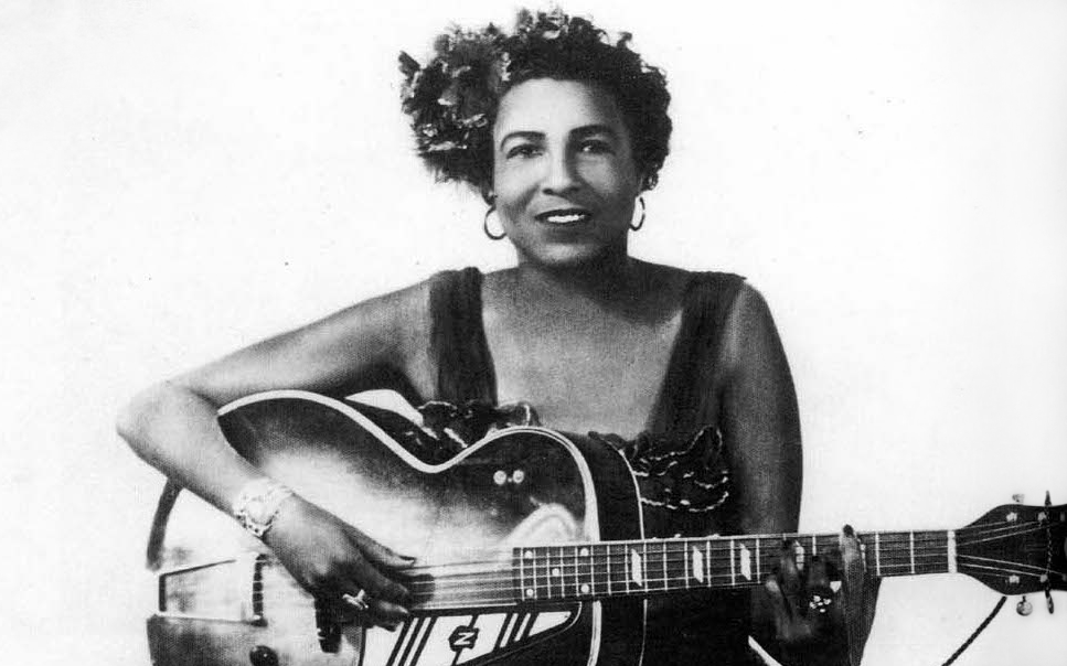 Blues music innovator Memphis Minnie is sitting down, holding her guitar, and smiling.