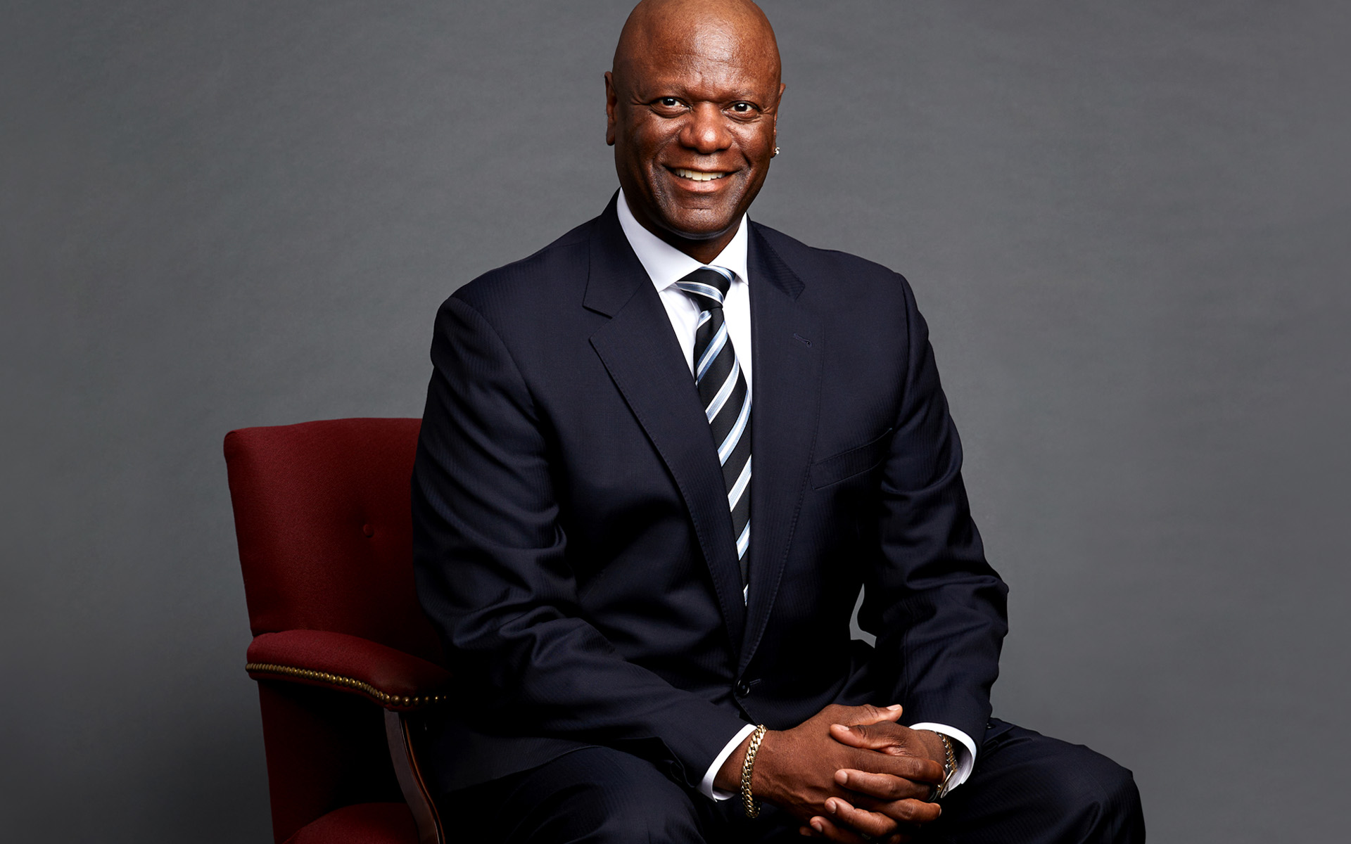 Prince Charles Alexander, Berklee Online instructor and vocal production engineer, is pictured sitting on a maroon chair, against a gray background.