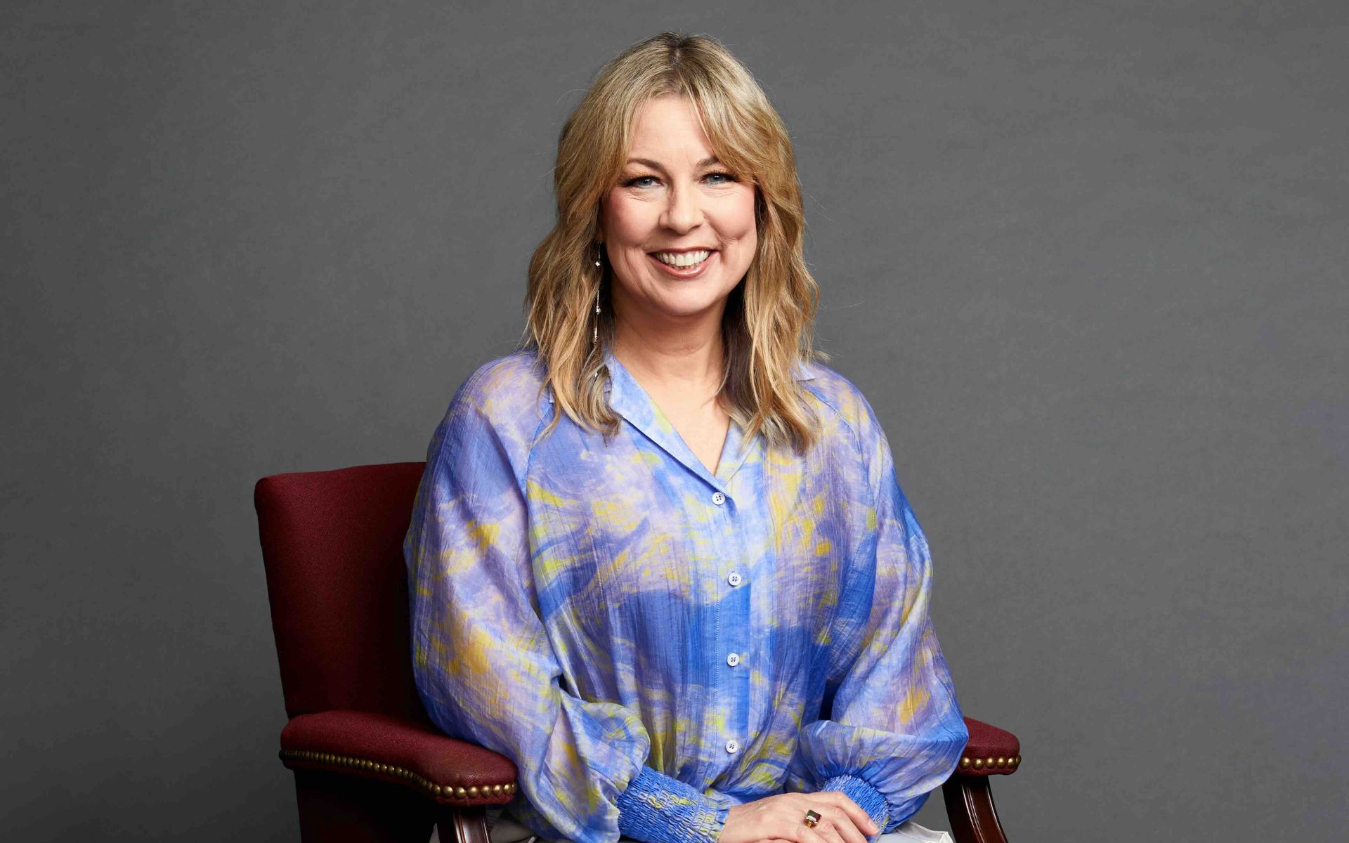 Adriana Balic is pictured, seated in a maroon chair, against a deep gray background. She is smiling and wearing a colorful blue shirt with purple hues.