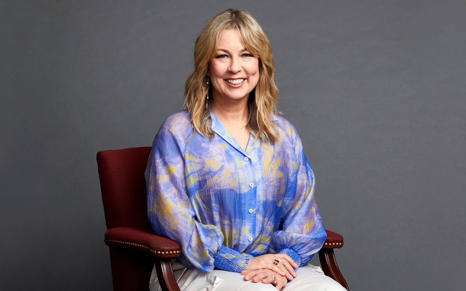 Adriana Balic is pictured, seated in a maroon chair, against a deep gray background. She is smiling and wearing a colorful blue shirt with purple hues.