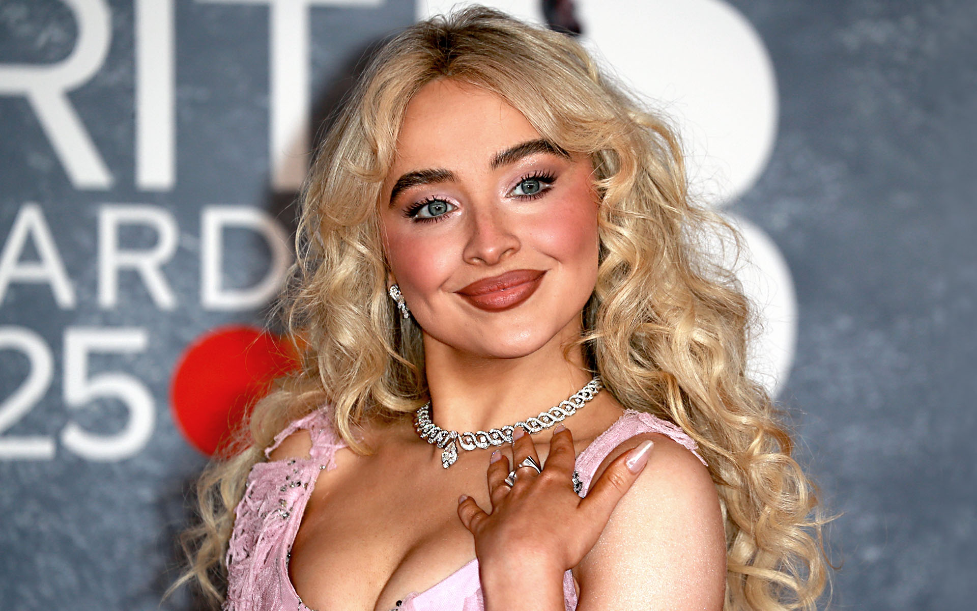 Sabrina Carpenter is pictured at the Intercontinental Hotel at The O2 in London, England, attending the BRIT Awards in March of 2025, wearing a pink dress and smiling.