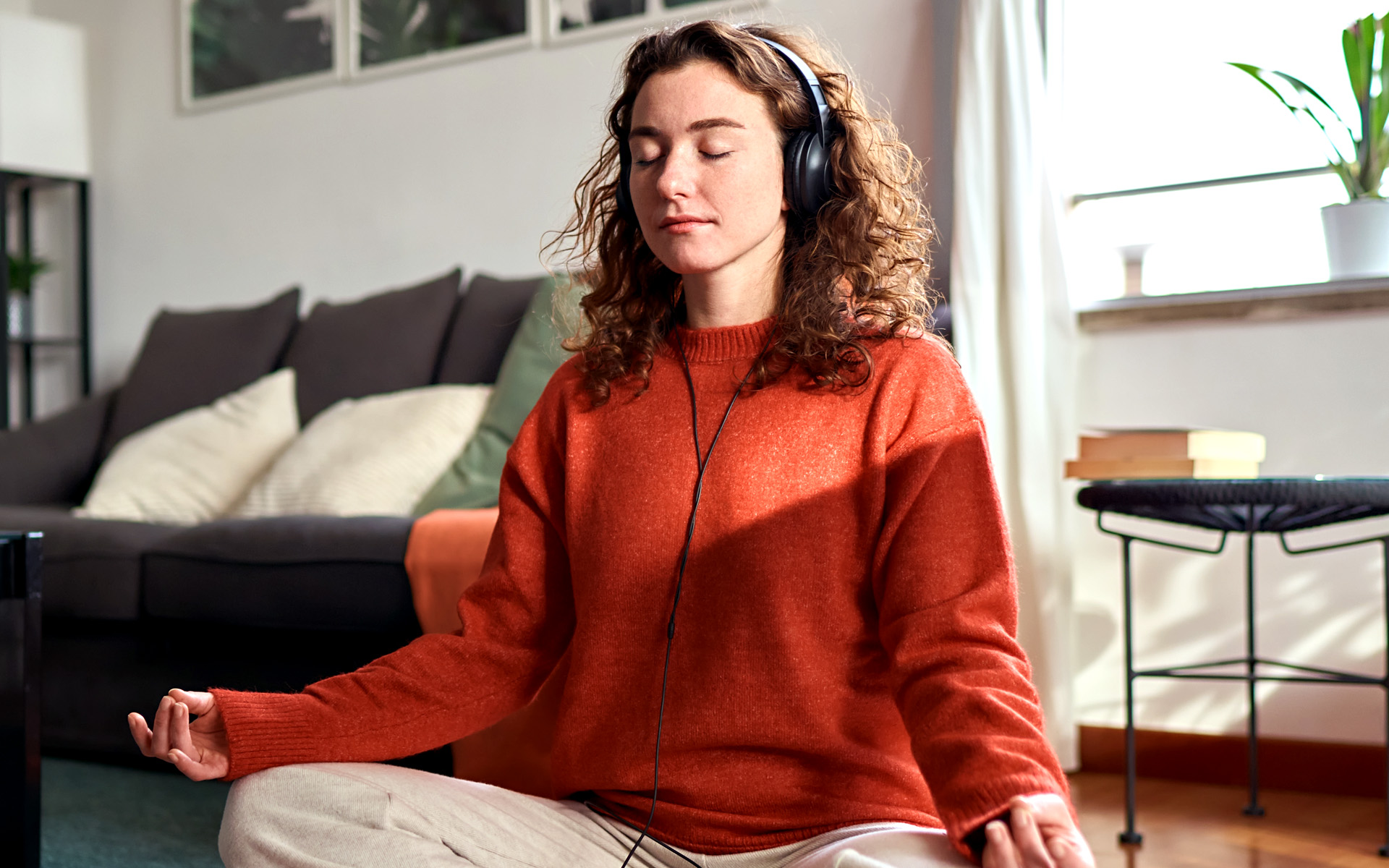 Person in an orange sweater sitting on the floor of their living room in a meditative pose with headphones on.