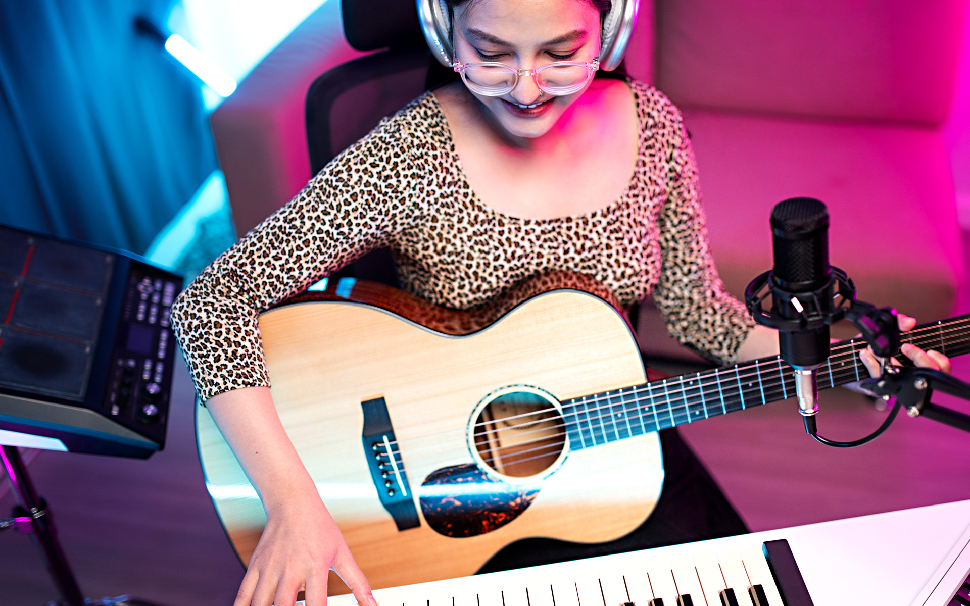 A teenage musician wears headphones and sits with a guitar in her lap in front of a computer with a MIDI keyboard.