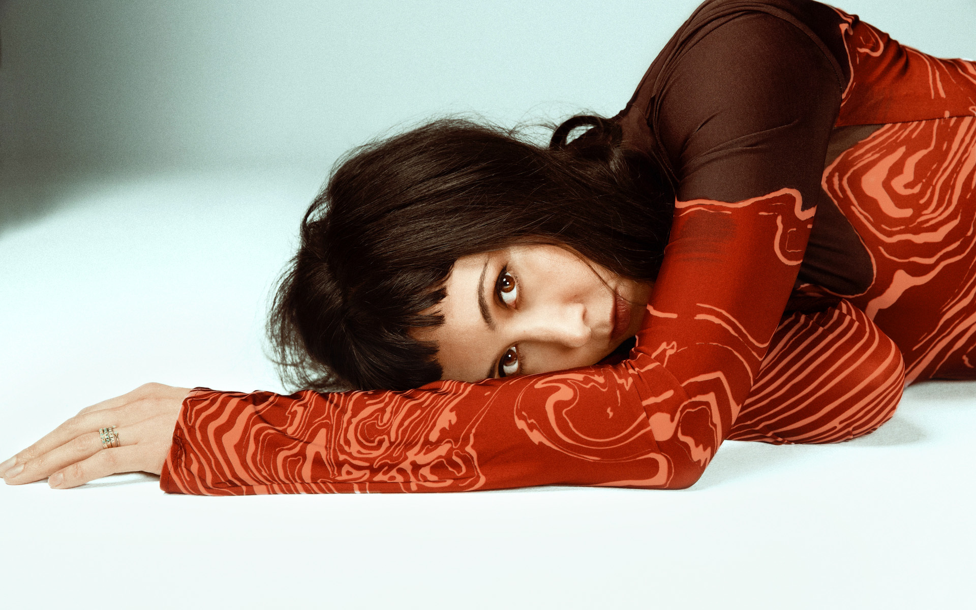 Vanessa Carlton lies on a white floor and stares at the camera, wearing a silky maroon shirt.