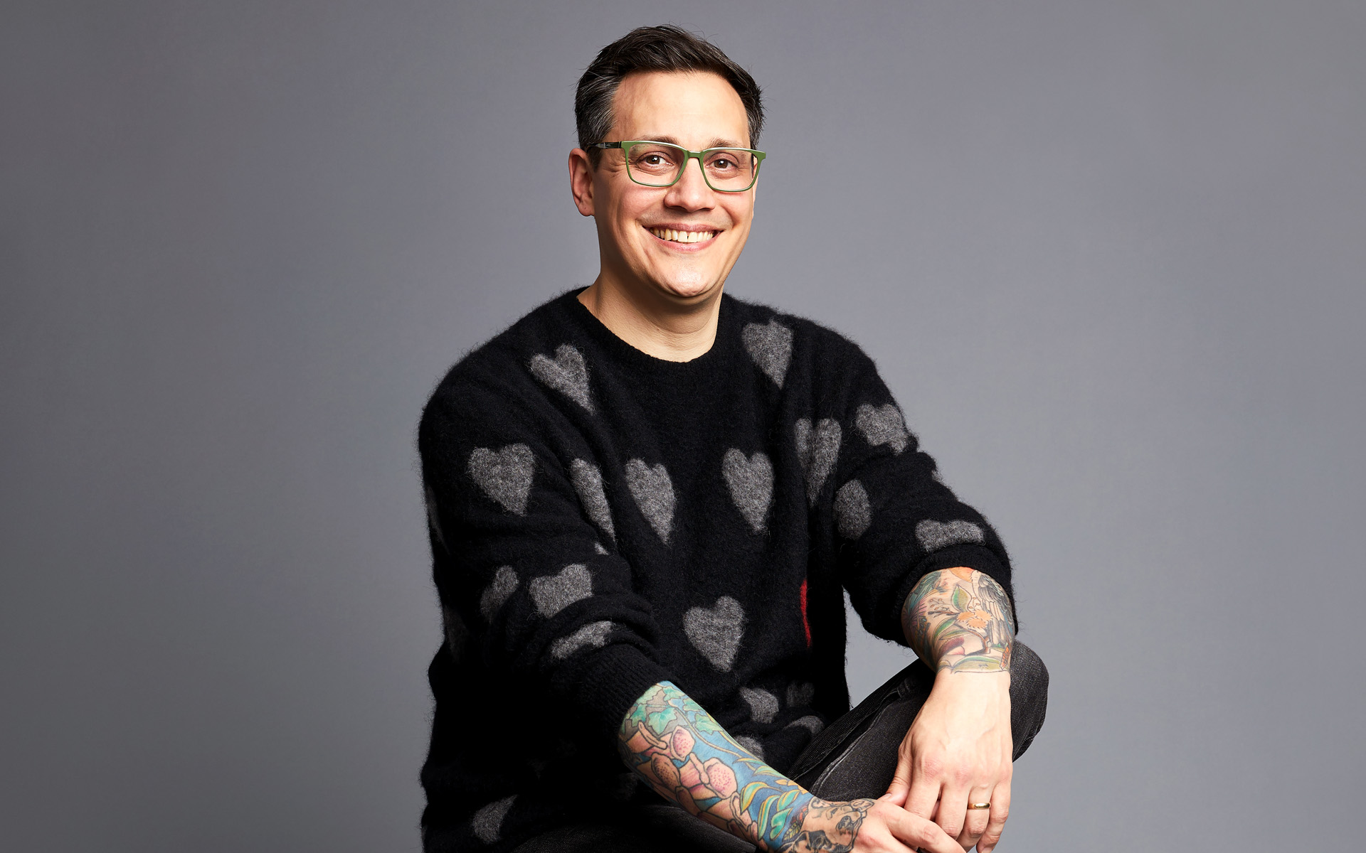 Owen Gurry sits against a gray background, wearing a black sweater with heart patterns. He has tattoos on both arms.