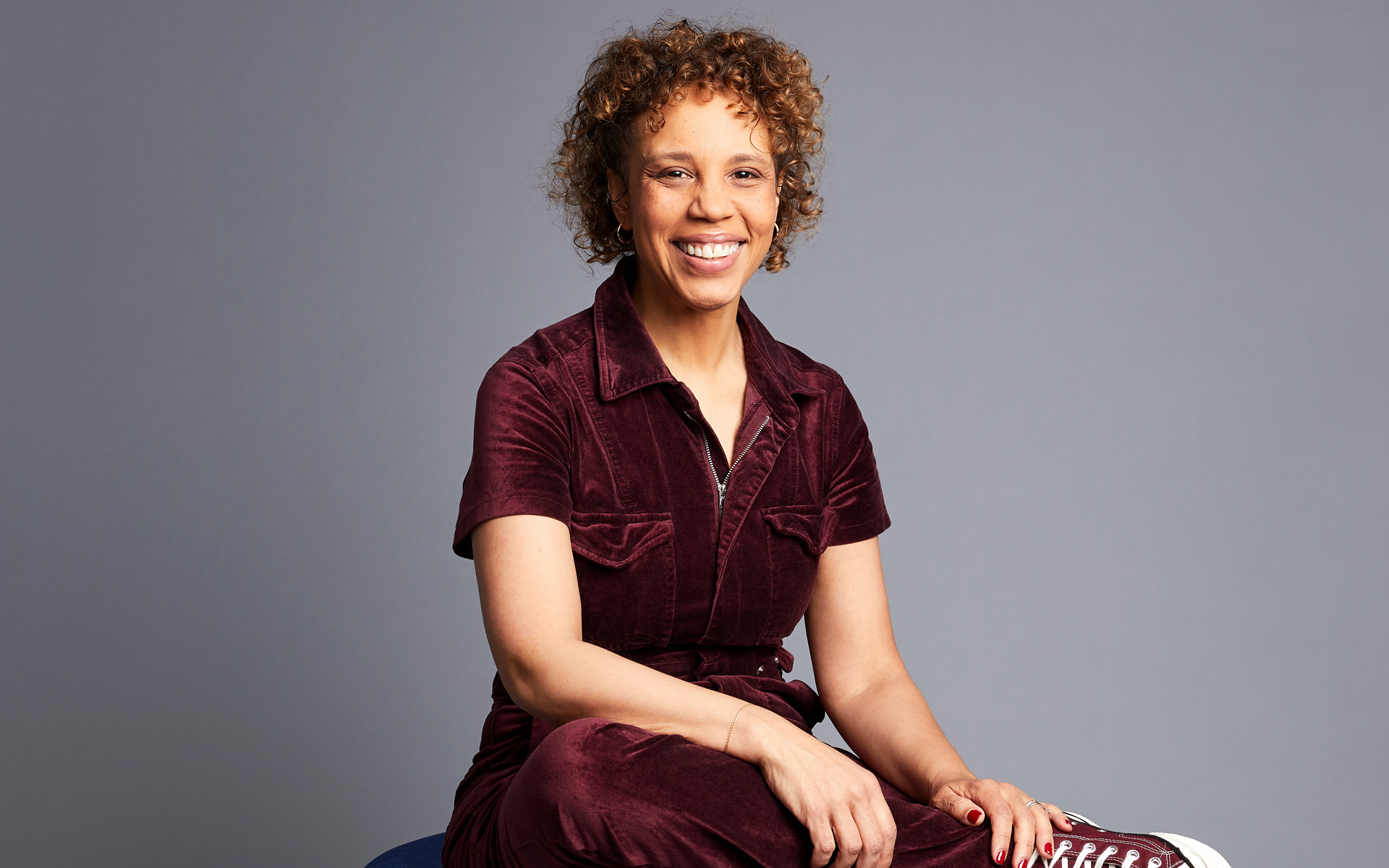 Shayna Steele sits against a gray background, wearing a maroon jumpsuit.