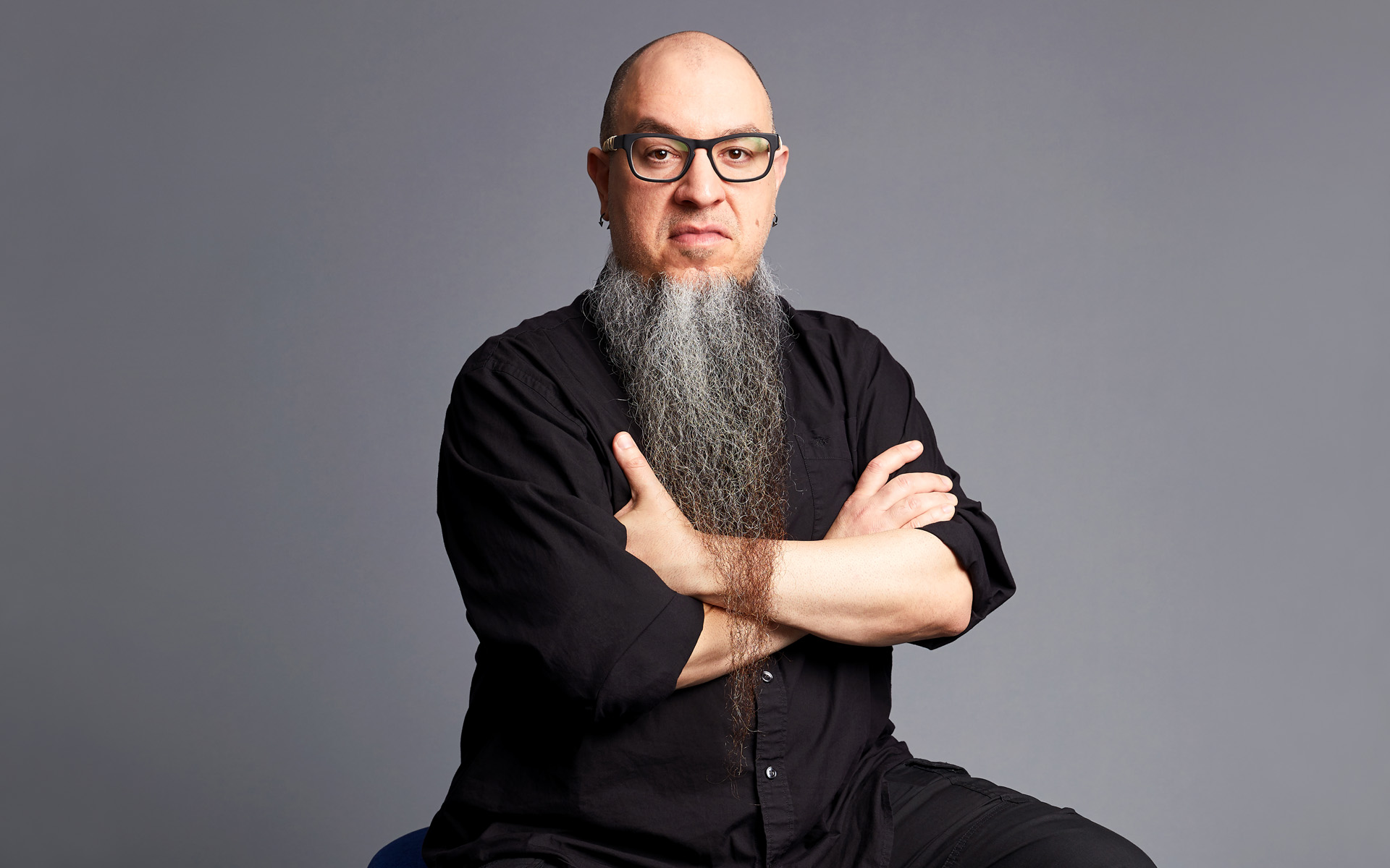 Enrique Gonzalez Müller sits against a gray background, wearing a black shirt and black pants. His beard is long and gray.
