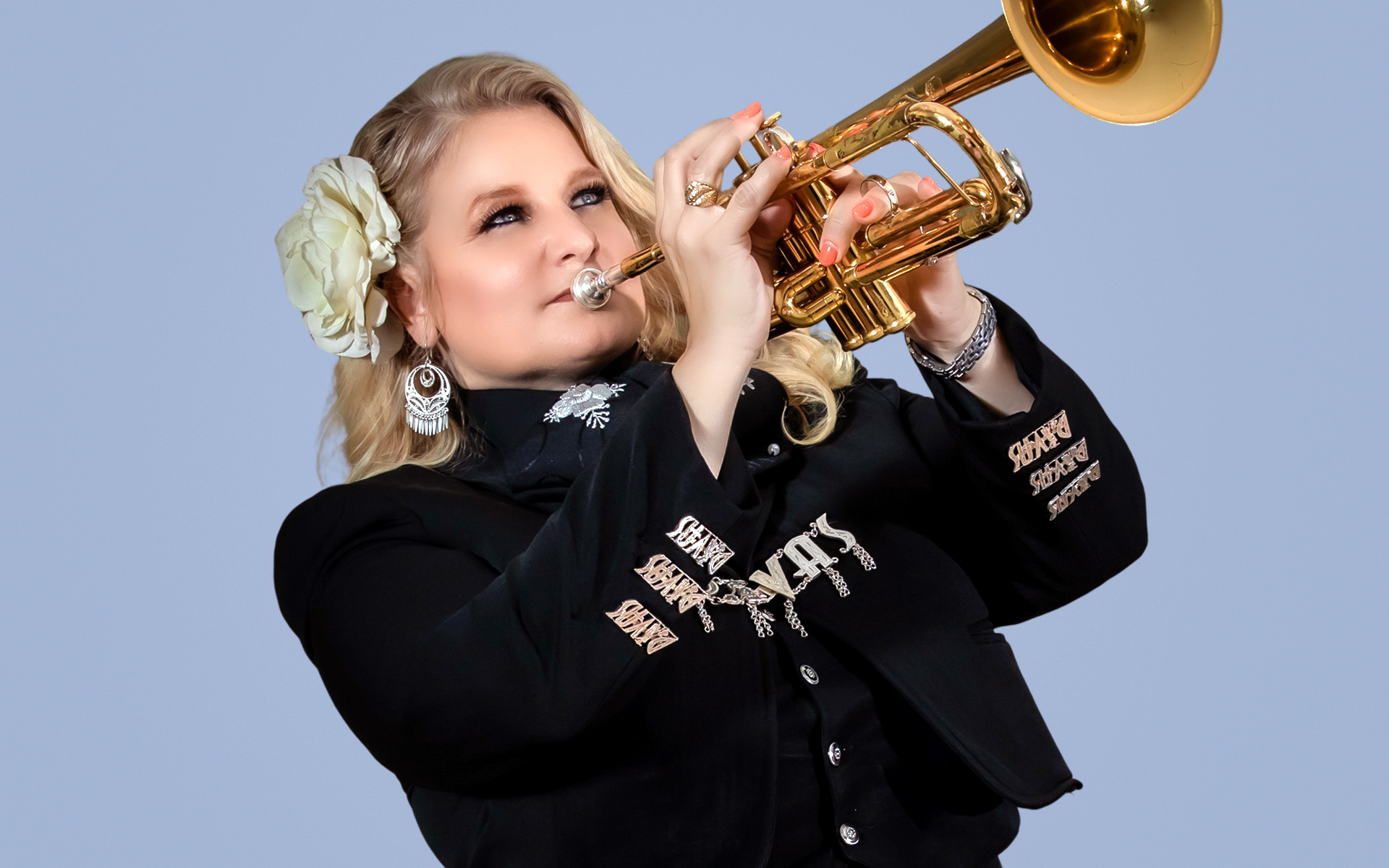Cindy Shea of Mariachi Divas wears a black jacket, a flower in her hair, and blows on her trumpet.