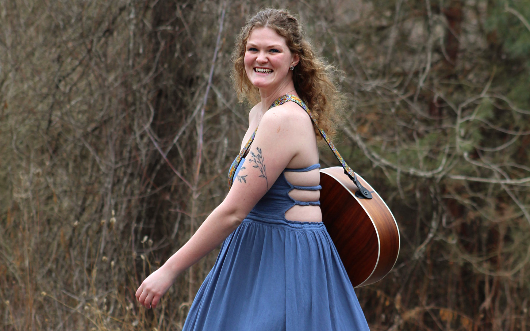 Hannah Crouch standing in the woods with her guitar wearing a blue dress