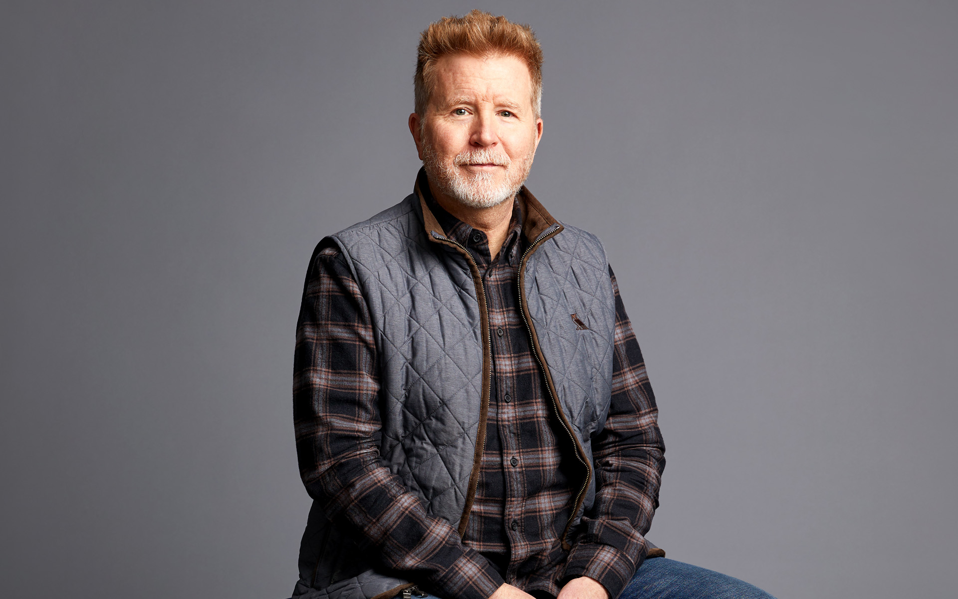Matt Rollings sits against a gray background, wearing a plaid shirt and a gray vest pants. His hair is short and red, and he has a gray beard.