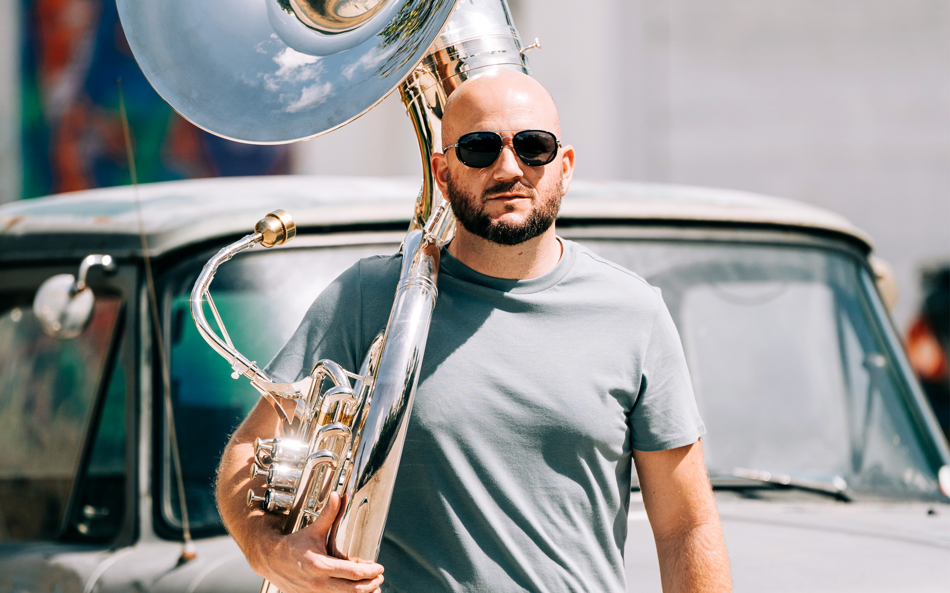 Bill Muter is pictured in this photograph, carrying his tuba and wearing a plain gray T-shirt, in front of a vintage automobile.