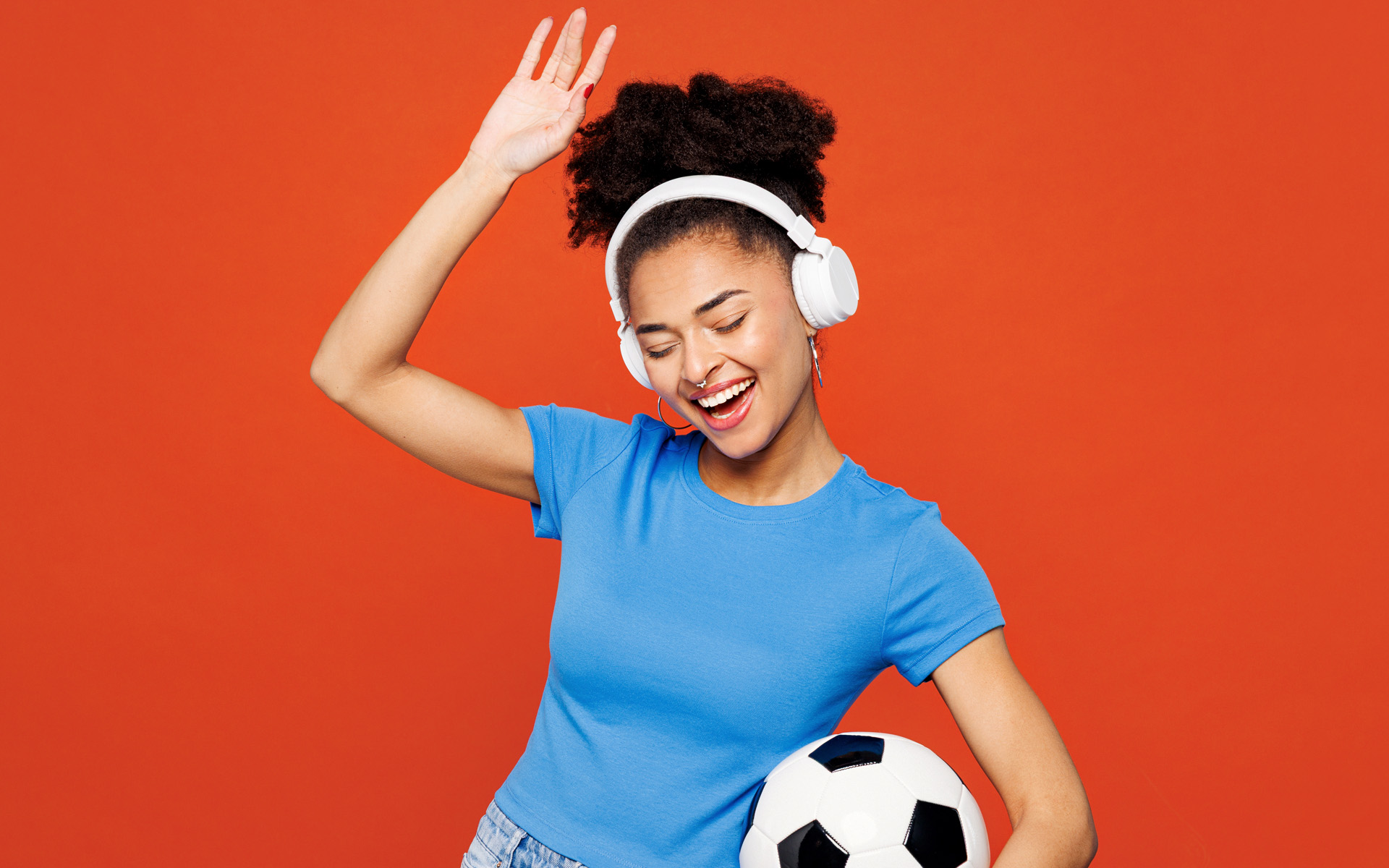 In this photograph, a woman in a blue shirt dances against a red background, waving one arm in the air, holding a soccer ball under the other arm, and wearing headphones on her head, symbolizing the music of the World Cup.