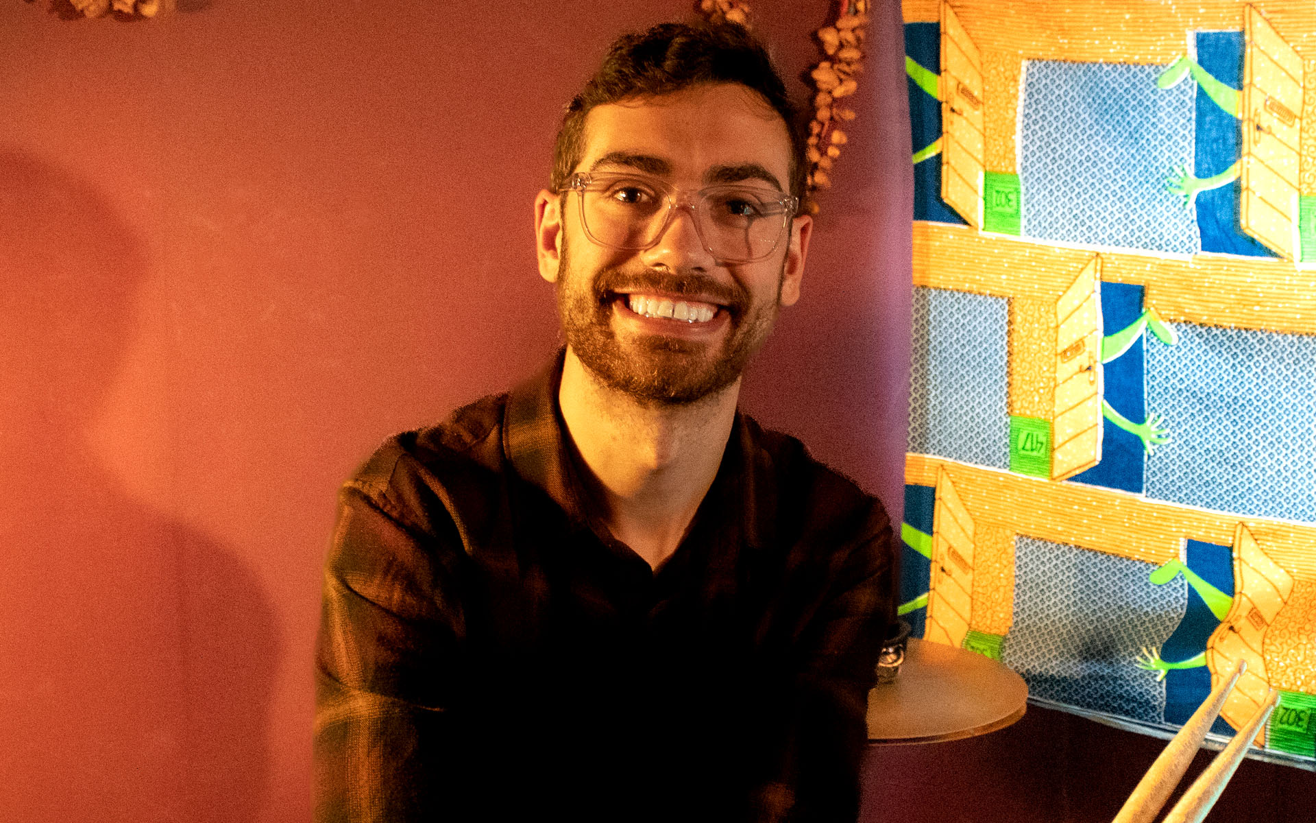 A picture of Kevin on a orange background, holding drum sticks.