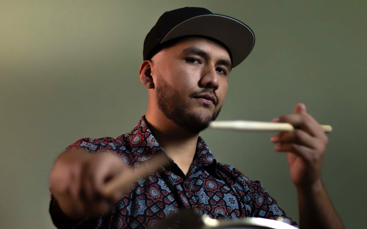 A picture of Oscar on a dark green background holding two drum sticks.