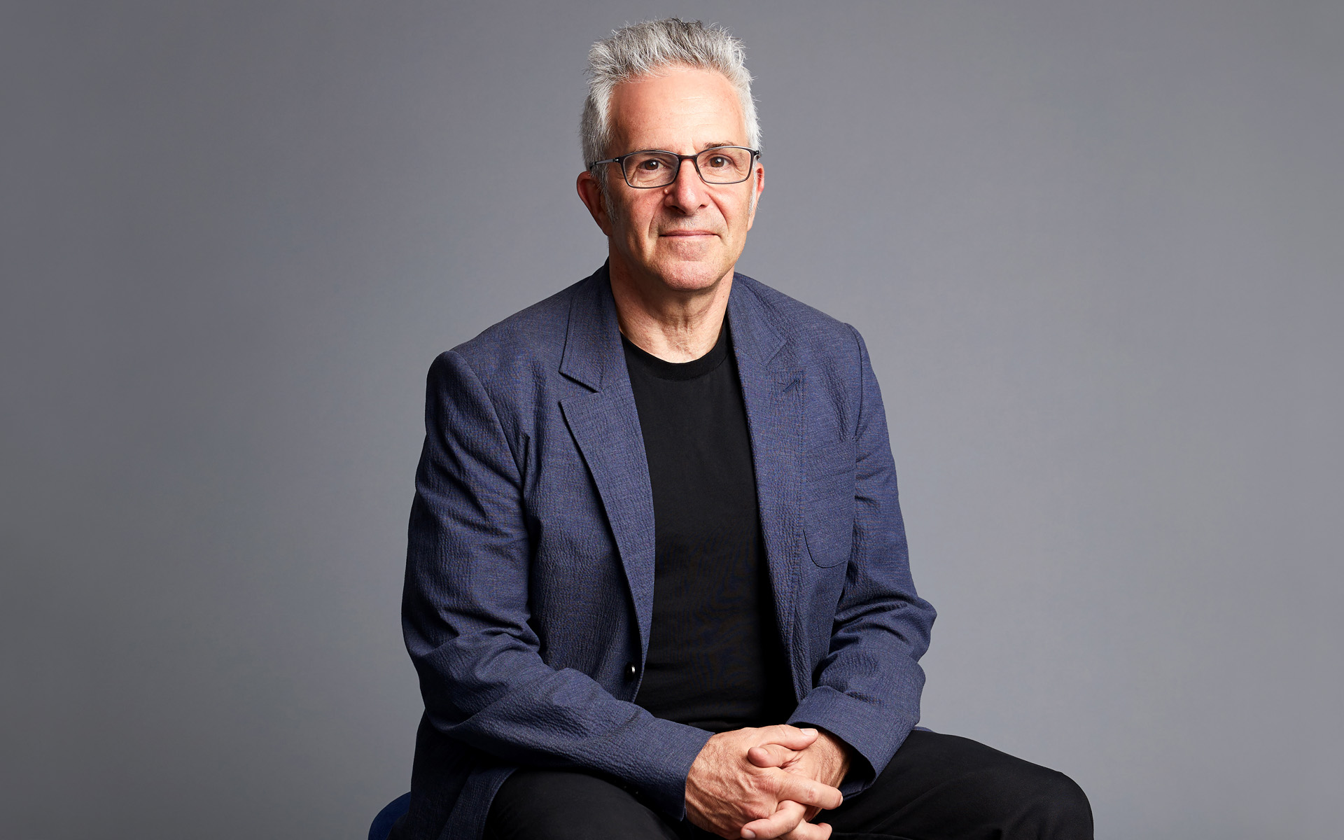 In this portrait, Jonathan Wyner sits against a gray background, wearing a black T-shirt and blue blazer. His gray hair is pushed back and his hands are clasped.