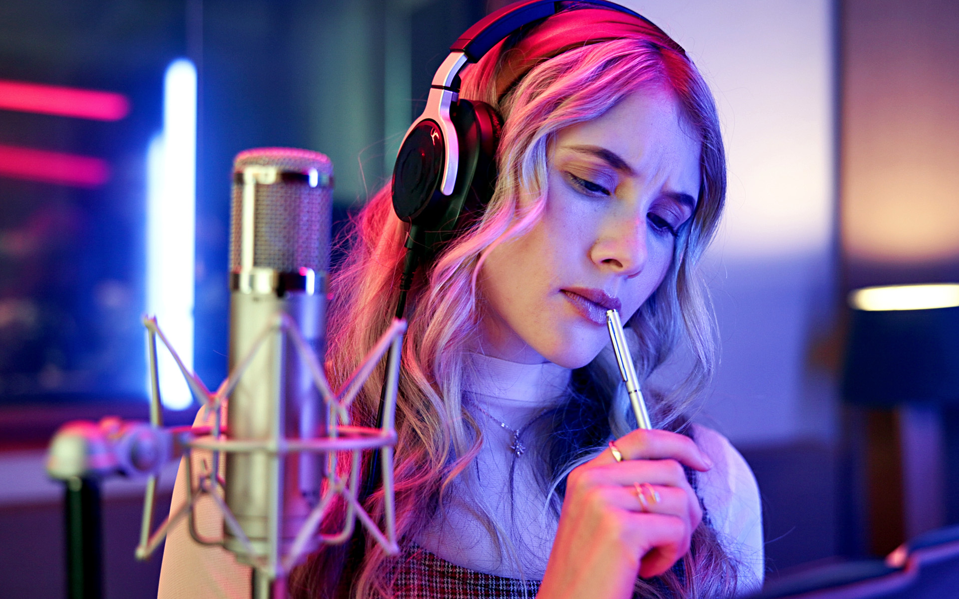 A songwriter in a recording studio who has a pen to their mouth trying to overcome writer's block.