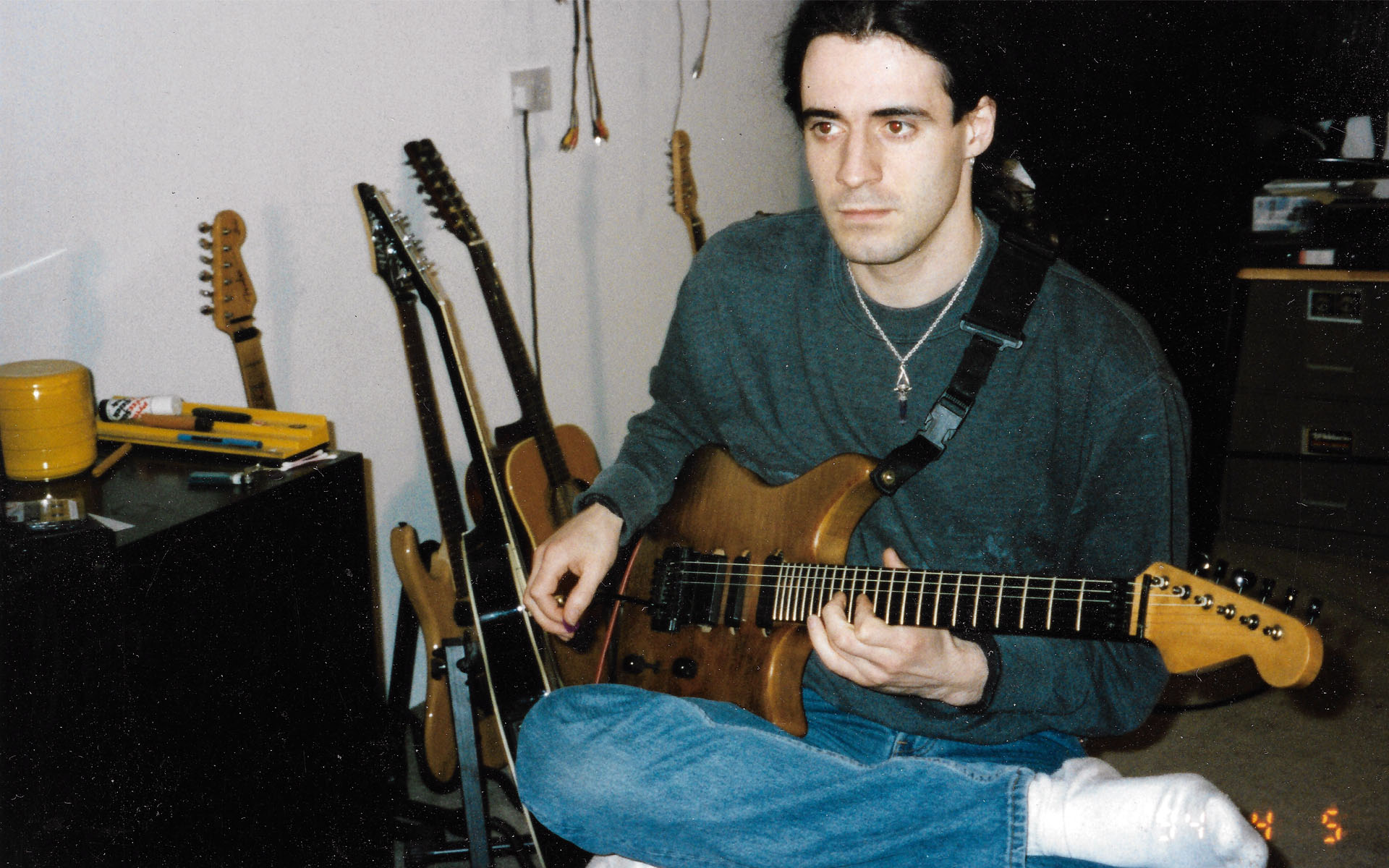 Guitarist Pete Langman is pictured in a photograph from the 1990s, sitting cross-legged on a floor, wearing bluejeans, white socks, a green sweater, and a necklace with a crystal on it.
