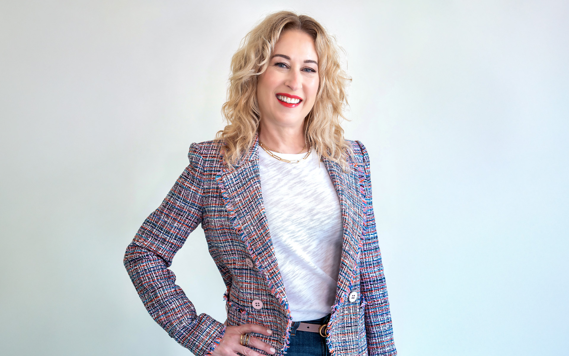 Portrait of Music Supervisor Julia Michels, standing against a white background and wearing a multi-colored blazer and blue jeans.