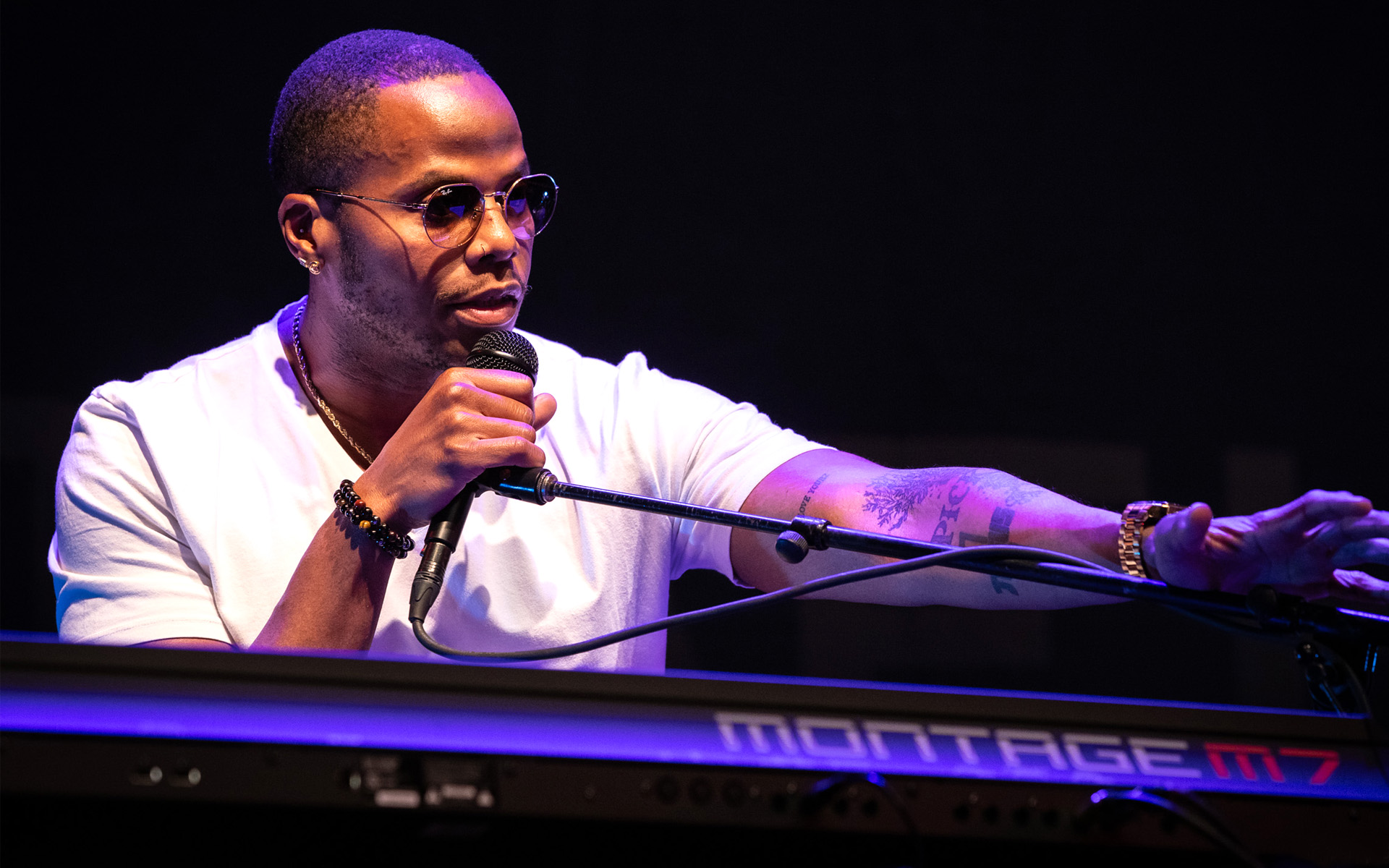 Tower of Power keyboardist Mike Jerel is pictured at his keys, wearing a white T-shirt and sunglasses, and reaching towards his microphone.